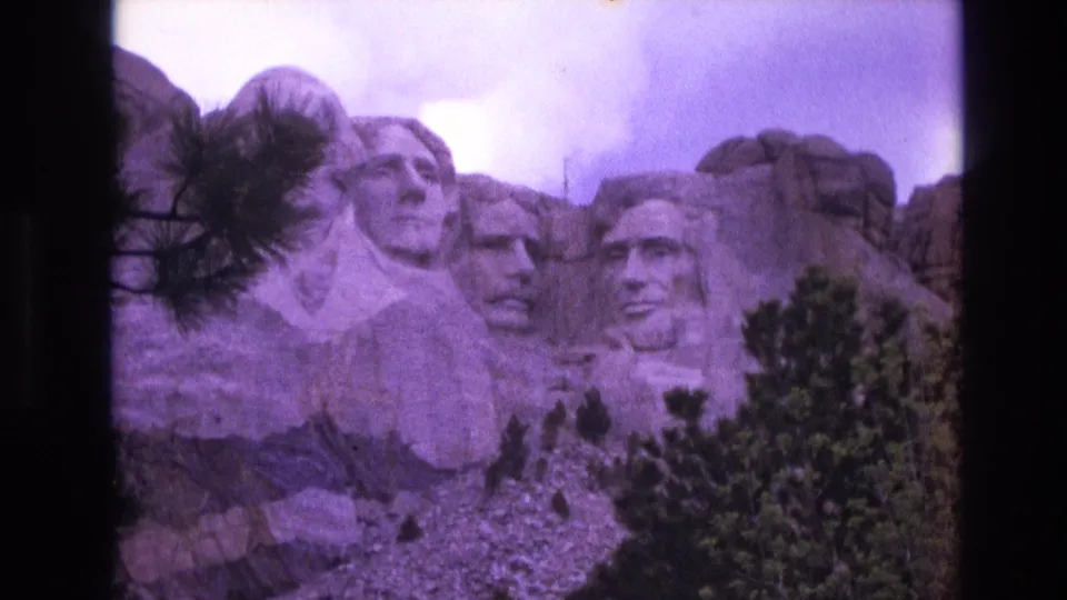 1974 South Dakota home-movie frame showing Mount Rushmore from a slightly shifted angle, with evergreen trees in the foreground and dark film borders at the sides.