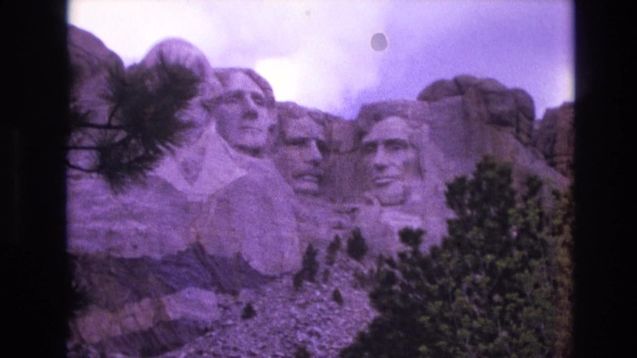 1974 8mm frame of Mount Rushmore in South Dakota, with the carved presidential heads seen through pine branches and a purple film color cast.