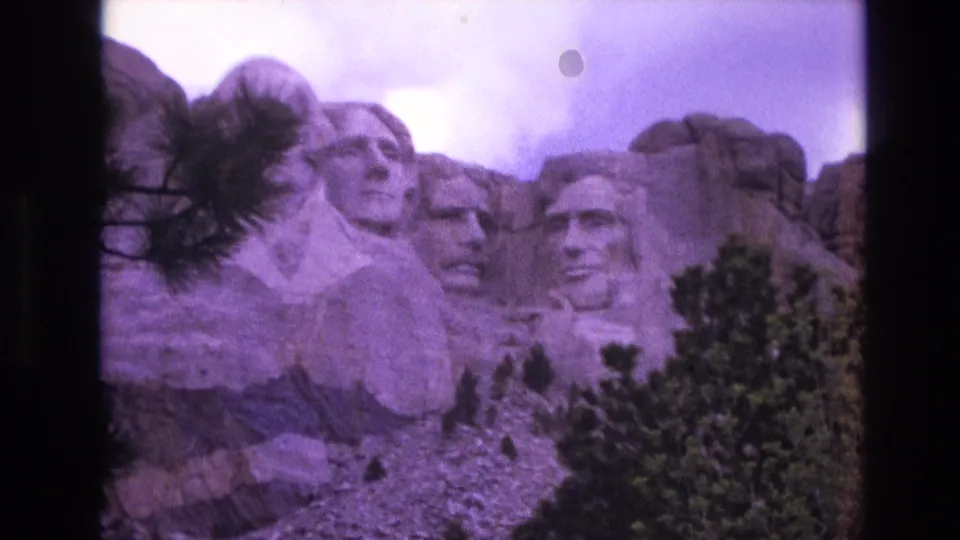 1974 8mm frame of Mount Rushmore in South Dakota, with the carved presidential heads seen through pine branches and a purple film color cast.