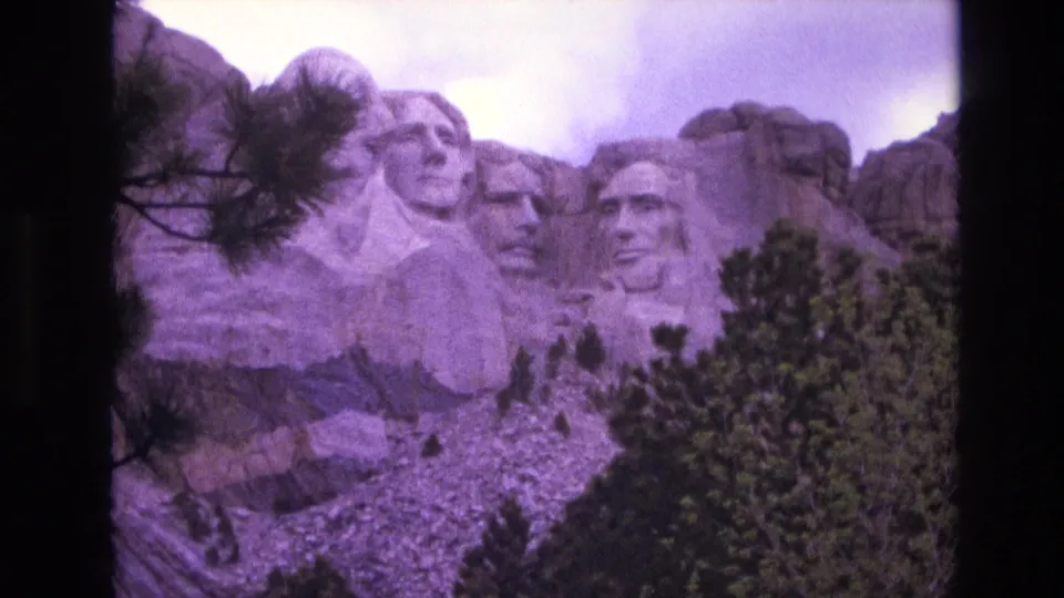 1974 8mm view of the Mount Rushmore presidential carvings above a rocky slope and pine trees, with visible grain and soft purple tint.
