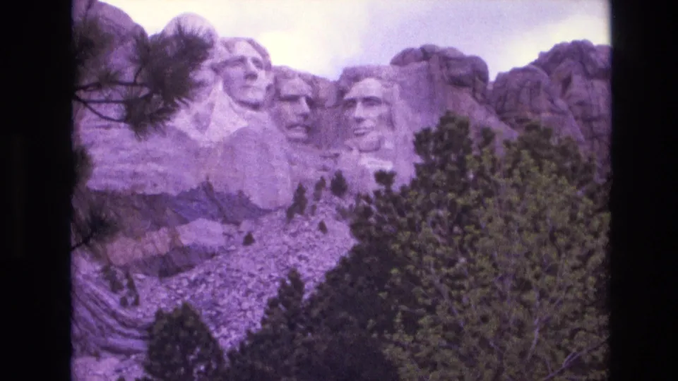 1974 South Dakota home-movie frame showing Mount Rushmore behind dense pines, with soft focus, grain, and dark film-frame edges.