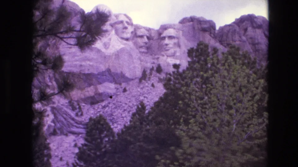 1974 8mm frame of Mount Rushmore with Washington, Jefferson, Roosevelt, and Lincoln visible across the granite face, partly framed by pine trees.