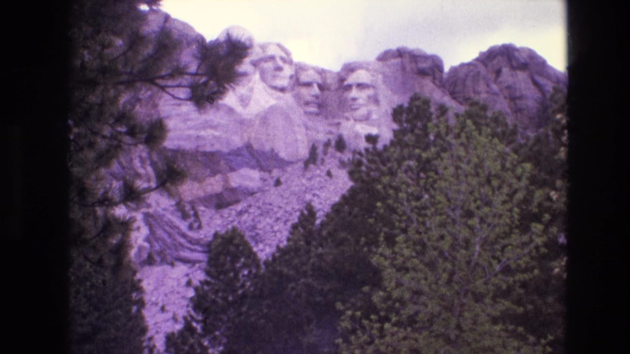 1974 8mm home-movie frame showing Mount Rushmore in South Dakota from a wider visitor vantage, with trees in the foreground and a purple-tinted film cast.