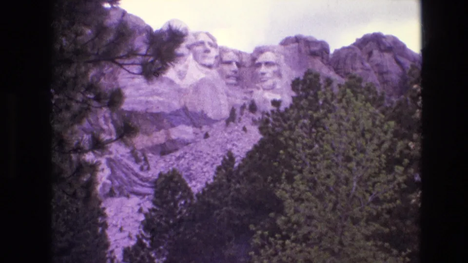 1974 8mm home-movie frame showing Mount Rushmore in South Dakota from a wider visitor vantage, with trees in the foreground and a purple-tinted film cast.