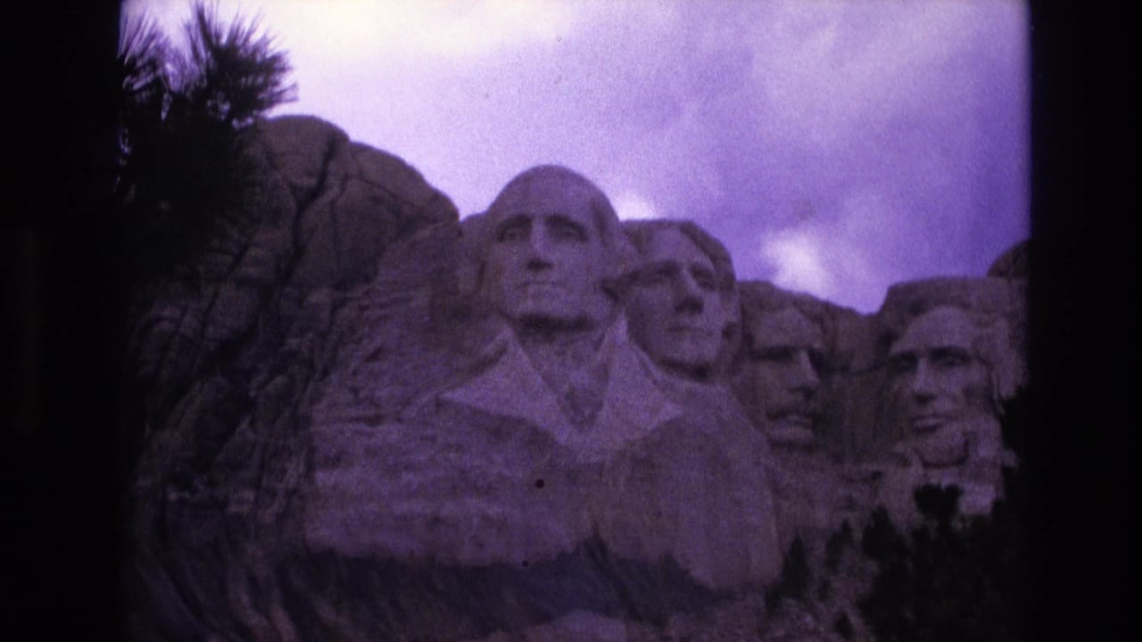 1974 8mm frame showing a closer visitor view of the Mount Rushmore carved presidential heads against granite and cloudy sky.