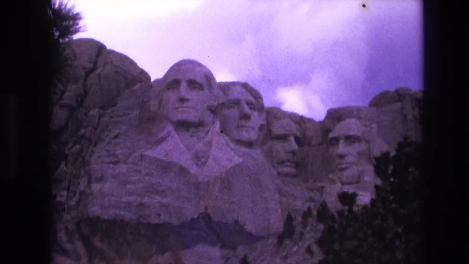 1974 South Dakota home-movie frame showing Mount Rushmore from a slightly shifted close angle, with carved faces, foreground trees, grain, and purple color fading.