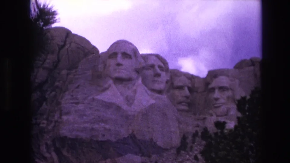 1974 8mm frame showing Mount Rushmore in South Dakota from a close visitor perspective, with trees below the carvings and a purple-tinted cloudy sky.