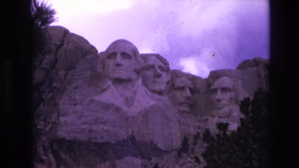 1974 8mm home-movie frame of Mount Rushmore in South Dakota, showing the presidential carvings below a cloudy sky with a purple film cast.