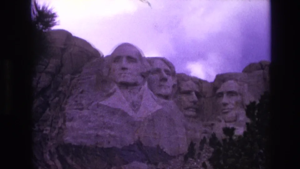 1974 South Dakota home-movie frame showing Mount Rushmore's presidential carvings, pine framing, cloudy sky, and visible 8mm film grain.