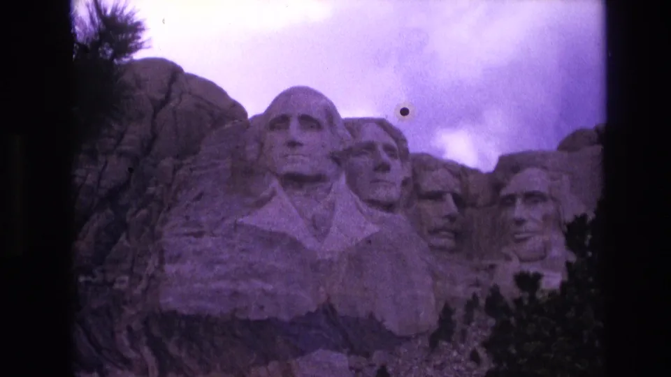1974 8mm frame of Mount Rushmore in South Dakota, with the monument below a purple-tinted cloudy sky and a noticeable dust mark in the scan.