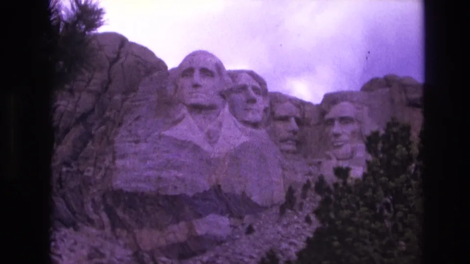 Final 1974 8mm home-movie frame of Mount Rushmore, showing the carved presidents, surrounding rock, lower trees, and cloudy sky in a purple color shift.
