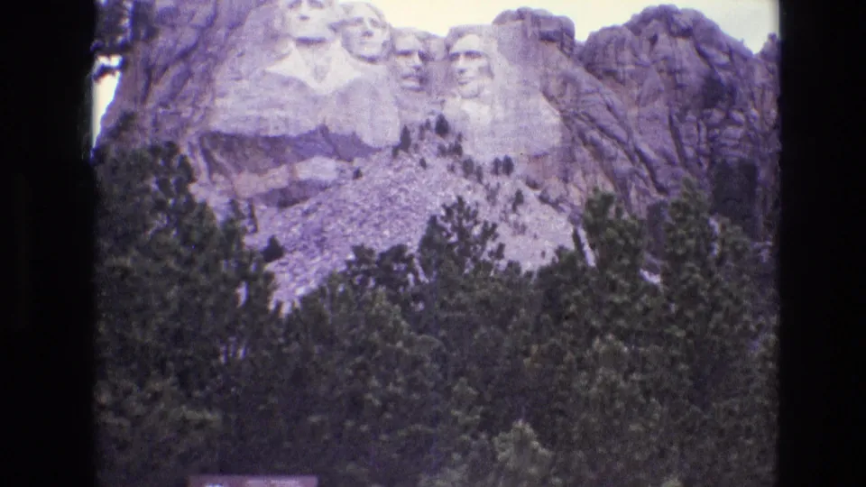 Wider 1974 home-movie view of Mount Rushmore National Memorial, with the monument rising above dense evergreens and visible 8mm frame borders.