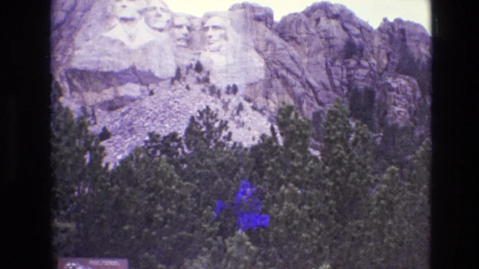 1974 8mm frame of Mount Rushmore in South Dakota behind evergreen foreground, showing aged color shift, soft detail, and a blue-purple stain near the lower right.