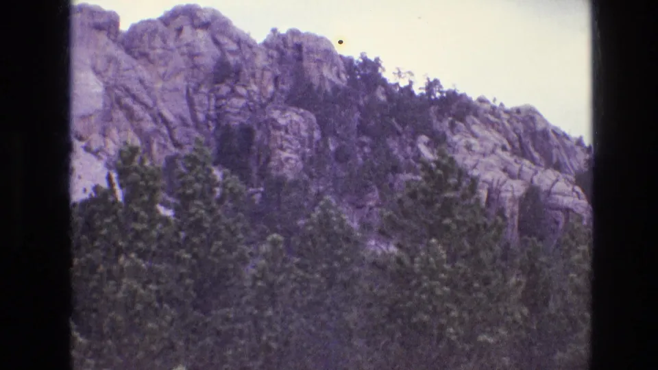 1974 8mm frame showing a partial view of Mount Rushmore at the left edge as the camera pans across pine trees and rugged granite outcrops.
