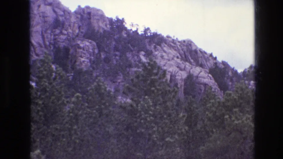 1974 8mm frame showing the rocky slope and evergreen trees around Mount Rushmore, with the presidential carving no longer clearly visible in this part of the pan.