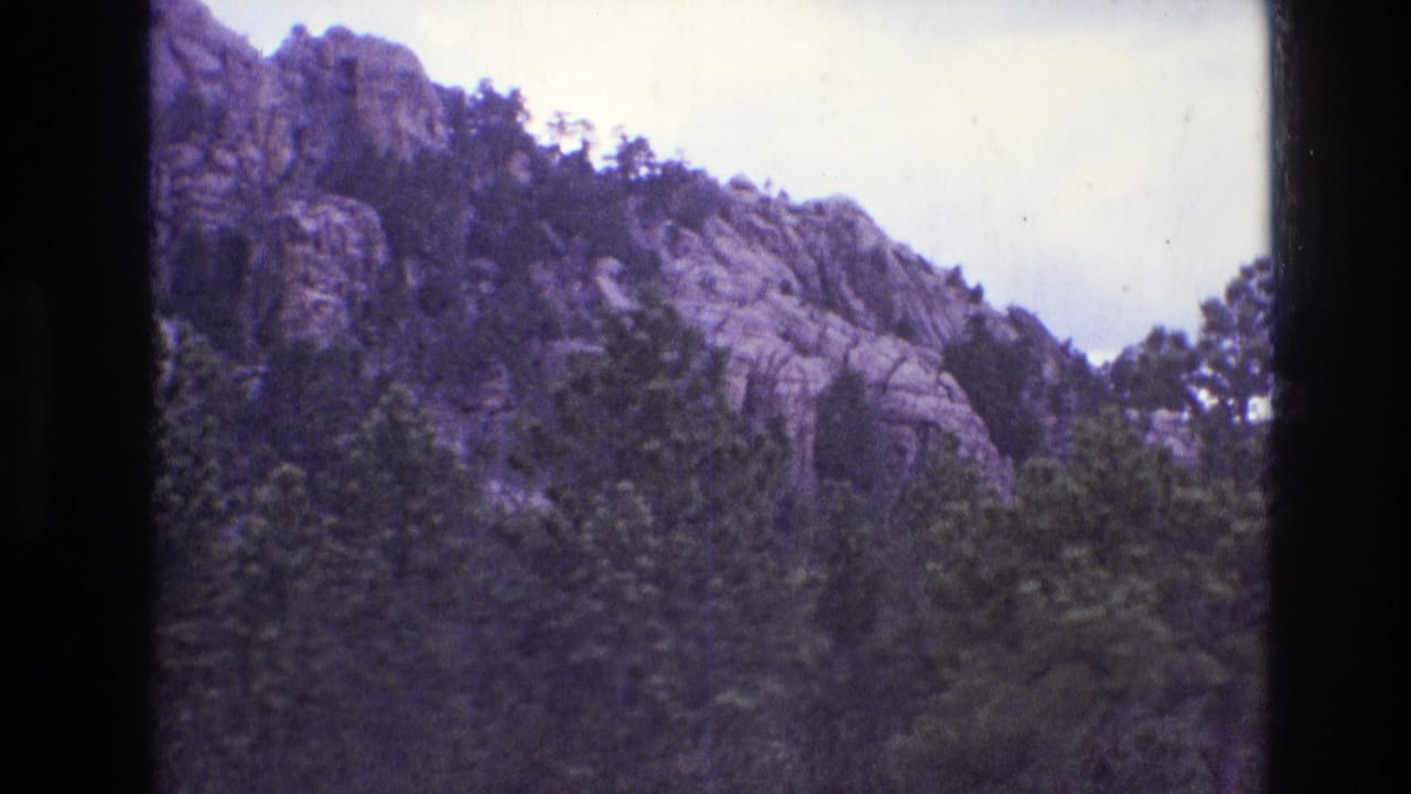 1974 South Dakota 8mm frame showing a rocky, tree-covered mountain slope beneath a cloudy sky with dark film borders and aged color cast.