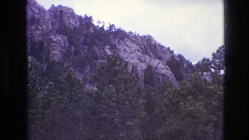 1974 South Dakota 8mm frame showing a rocky, tree-covered mountain slope beneath a cloudy sky with dark film borders and aged color cast.