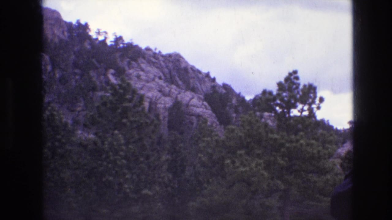 1974 South Dakota home-movie frame with a rugged rock face, dense evergreens, overcast sky, and the visible black edge of the 8mm frame.