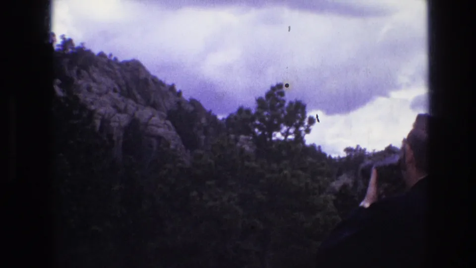 1974 South Dakota 8mm frame showing an adult visitor in dark clothing lifting a camera toward a rocky, forested mountainside beneath heavy clouds.