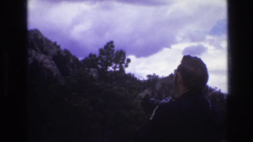 1974 South Dakota home-movie frame with an adult seen from behind at the lower right, facing a forested rocky hillside beneath a cloud-filled sky.