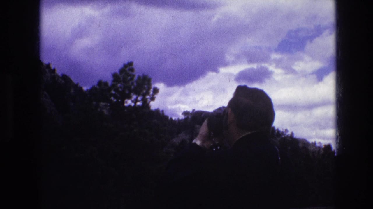 In 1974 South Dakota 8mm footage, a silhouetted adult man raises a camera toward a dramatic cloudy sky over a dark wooded ridge.