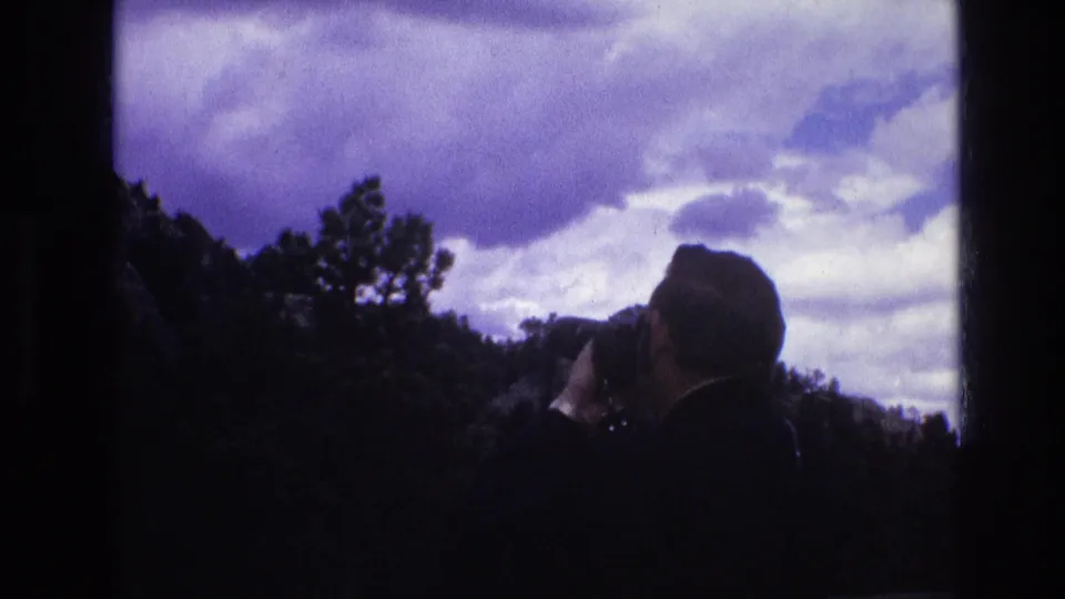 In 1974 South Dakota 8mm footage, a silhouetted adult man raises a camera toward a dramatic cloudy sky over a dark wooded ridge.