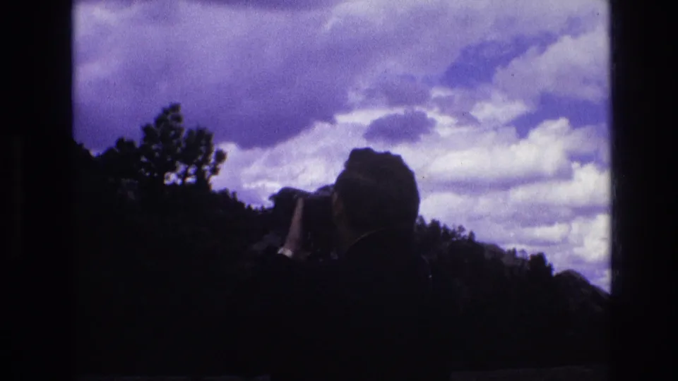 The silhouetted man remains in profile with a camera raised, framed against a purple-blue sky and dark South Dakota ridge in the supplied 1974 home-movie clip.