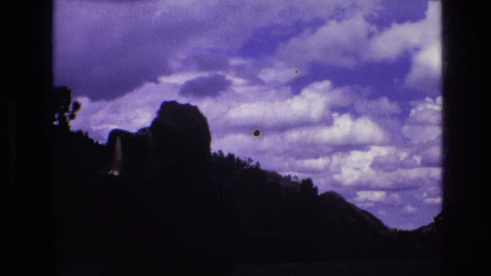 A mostly silhouetted view shows the man's head and camera near the lower frame edge, with dark hills and a broad cloud-filled sky in 1974 South Dakota home-movie footage.