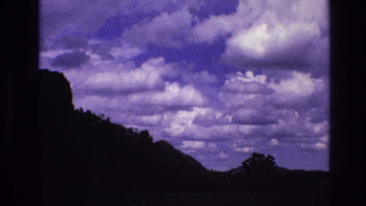 1974 South Dakota 8mm frame showing a cloud-filled purple-blue sky over a dark silhouetted hillside, tree line, and low rural horizon.