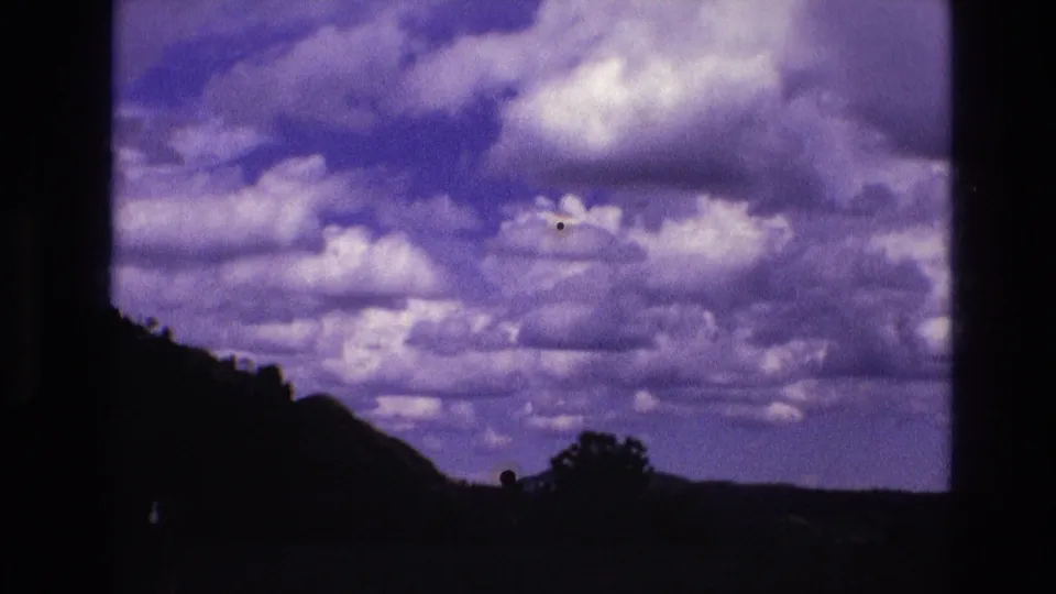 1974 South Dakota 8mm frame showing large clouds over a shadowed hillside, low horizon, and central tree silhouette with soft film blur.