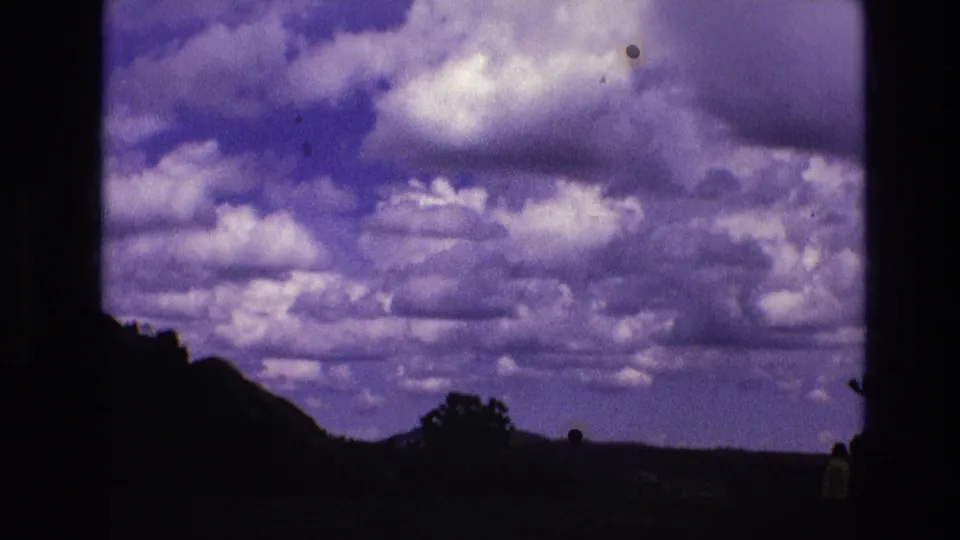 1974 South Dakota 8mm frame of a cloud-heavy purple-blue sky above silhouetted hills and trees, with small dark film specks near the frame.