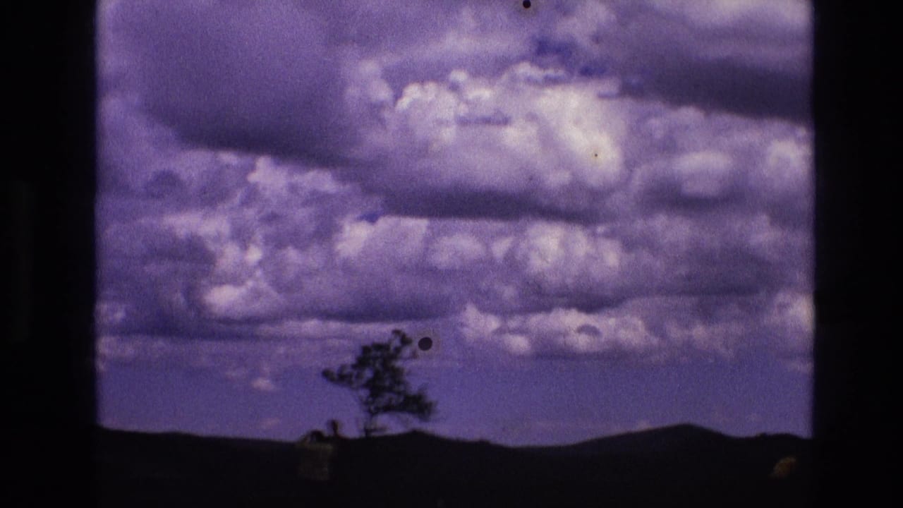 1974 South Dakota home-movie frame showing purple-tinged clouds above dark rolling hills, with a lone tree and small foreground silhouettes near Mount Rushmore National Memorial.