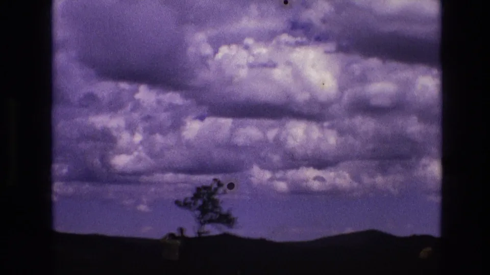1974 South Dakota home-movie frame showing purple-tinged clouds above dark rolling hills, with a lone tree and small foreground silhouettes near Mount Rushmore National Memorial.