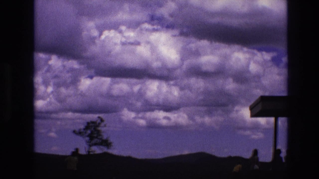 Cloudy sky over dark hills with a lone tree, small visitors, and an overlook canopy at right.