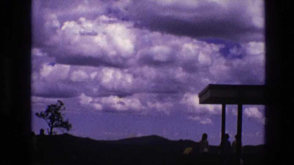 1974 South Dakota home-movie frame showing a cloud-heavy sky, dark hillside, lone tree, and silhouetted visitors standing near an overlook canopy by Mount Rushmore National Memorial.
