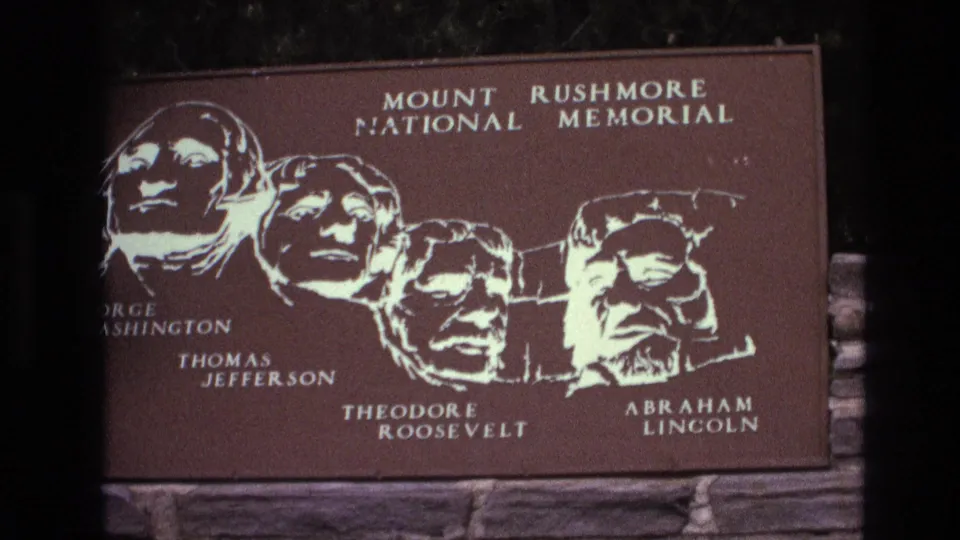 1974 South Dakota home-movie frame showing a Mount Rushmore National Memorial sign with illustrated portraits labeled for George Washington, Thomas Jefferson, Theodore Roosevelt, and Abraham Lincoln.