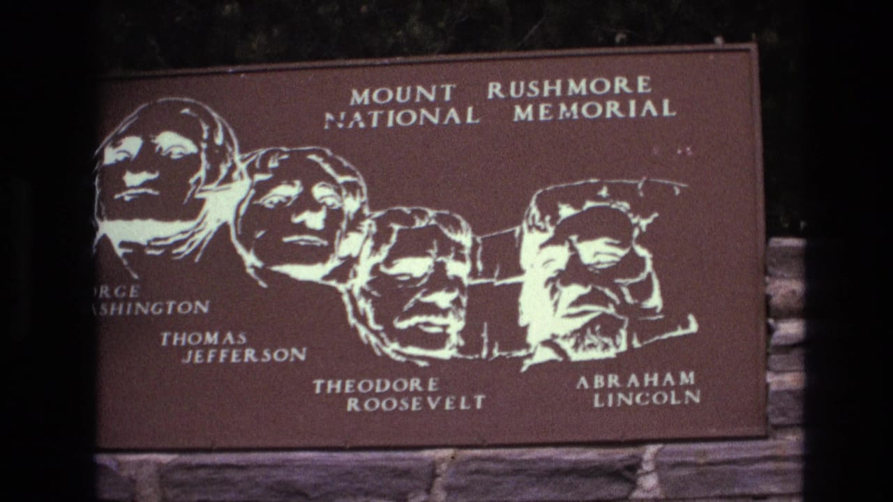 1974 South Dakota 8mm frame showing a brown Mount Rushmore National Memorial plaque with white portraits and names of Washington, Jefferson, Roosevelt, and Lincoln above a stone wall.