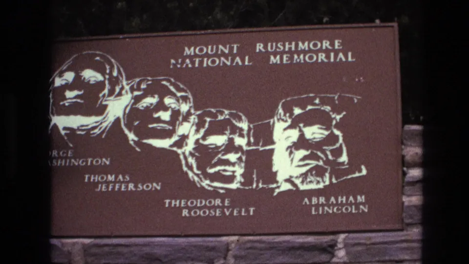 1974 South Dakota 8mm frame showing a brown Mount Rushmore National Memorial plaque with white portraits and names of Washington, Jefferson, Roosevelt, and Lincoln above a stone wall.