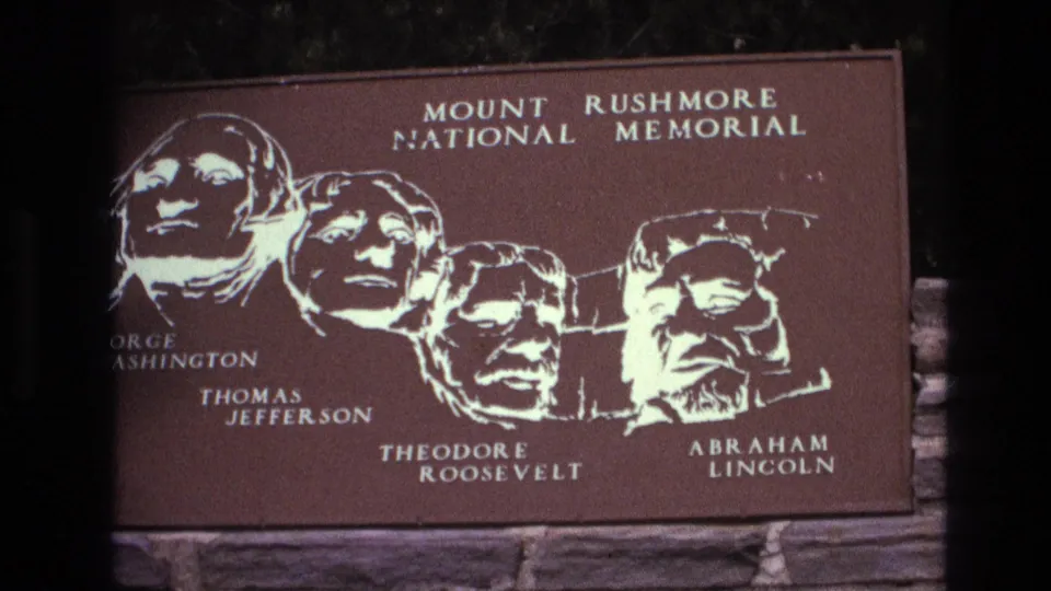 1974 South Dakota 8mm frame of the Mount Rushmore National Memorial sign, with the four presidential portrait silhouettes and names visible against a brown plaque surface.
