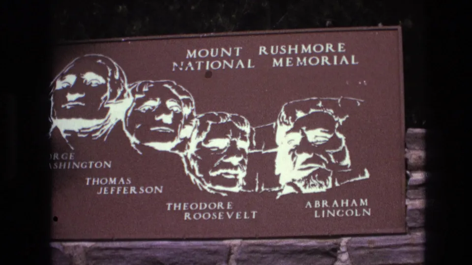 1974 South Dakota 8mm frame showing the Mount Rushmore National Memorial plaque from a slightly lower angle, with the presidential names and white portrait outlines visible.
