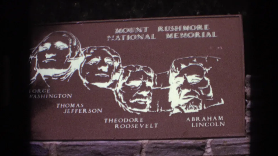 1974 South Dakota 8mm frame showing a slightly tilted visitor view of the Mount Rushmore National Memorial plaque, with presidential silhouettes, names, and the stone mounting wall visible.