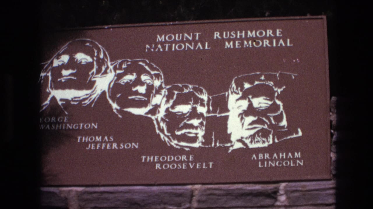 1974 South Dakota 8mm frame showing a close view of a Mount Rushmore National Memorial sign with illustrated heads of Washington, Jefferson, Theodore Roosevelt, and Lincoln.