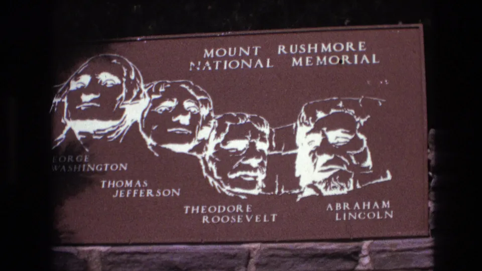 1974 8mm home-movie frame of the Mount Rushmore National Memorial sign in South Dakota, with the four named presidents rendered in white on a brown plaque.