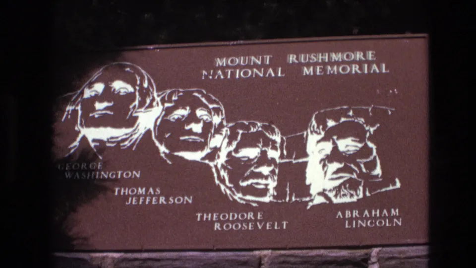 1974 South Dakota visitor footage frame showing the Mount Rushmore National Memorial sign with white portrait artwork and labels for the four presidents.