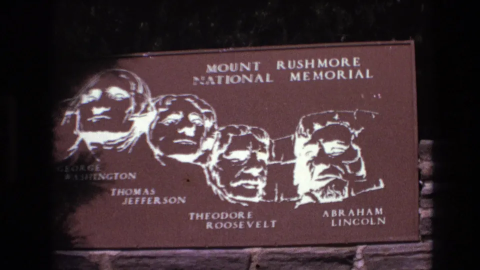 Final 1974 8mm frame of the Mount Rushmore National Memorial sign in South Dakota, with the four presidential illustrations and names visible in soft low light.
