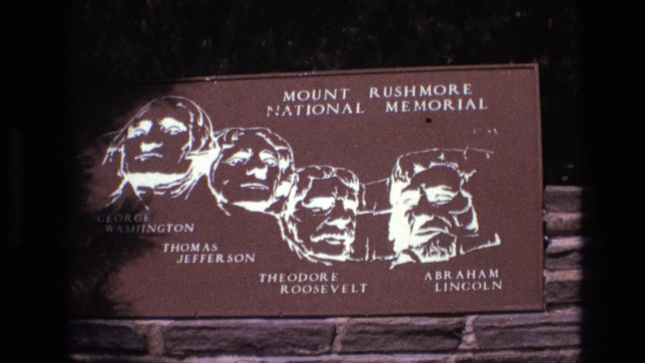 1974 8mm frame from South Dakota showing a Mount Rushmore National Memorial plaque with the carved presidential heads labeled George Washington, Thomas Jefferson, Theodore Roosevelt, and Abraham Lincoln.