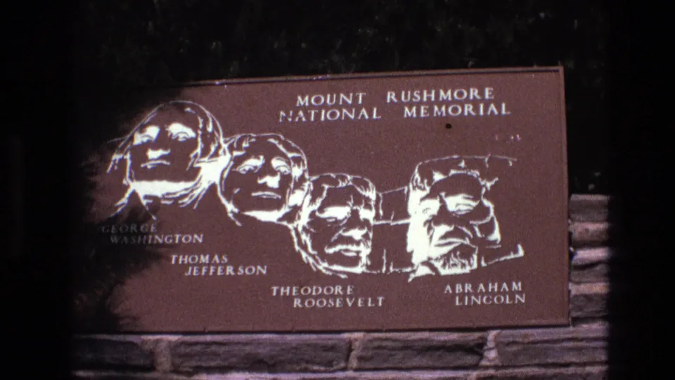 1974 8mm frame from South Dakota showing a Mount Rushmore National Memorial plaque with the carved presidential heads labeled George Washington, Thomas Jefferson, Theodore Roosevelt, and Abraham Lincoln.