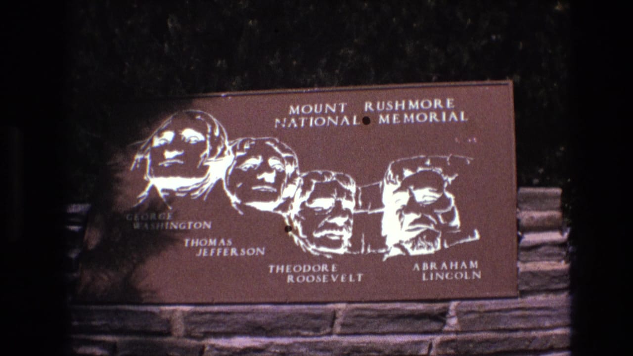 1974 8mm frame at Mount Rushmore National Memorial showing the bronze-toned presidential plaque with a red-brown elevation sign entering the lower frame.