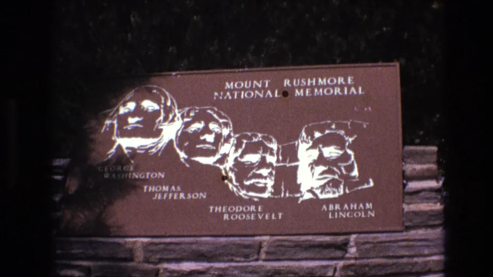 1974 8mm frame at Mount Rushmore National Memorial showing the bronze-toned presidential plaque with a red-brown elevation sign entering the lower frame.