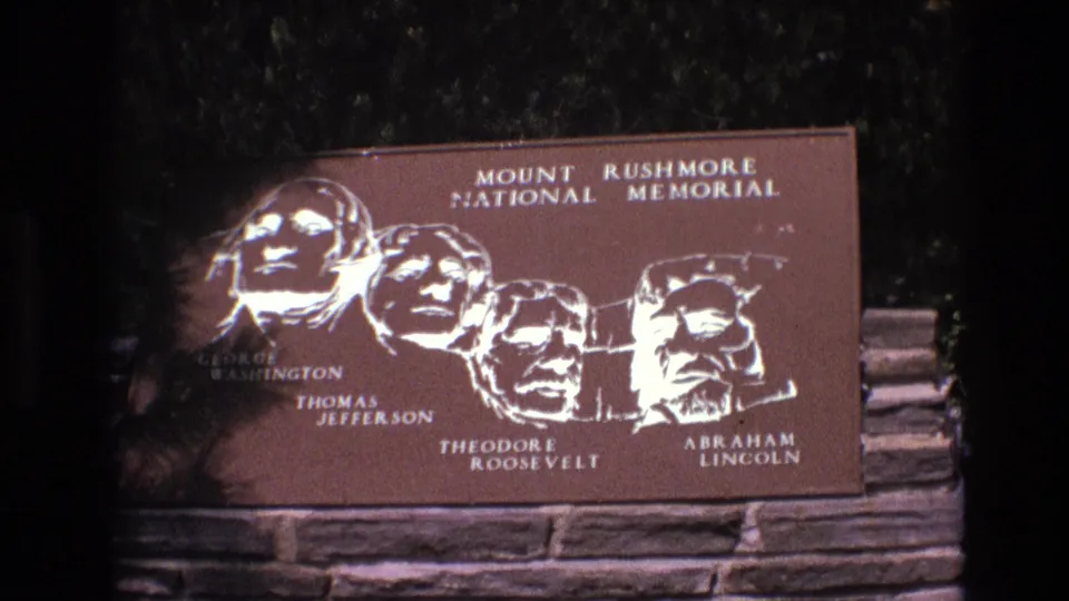 1974 8mm frame showing a close visitor-shot view of the Mount Rushmore National Memorial plaque and its labeled depictions of the four presidents.